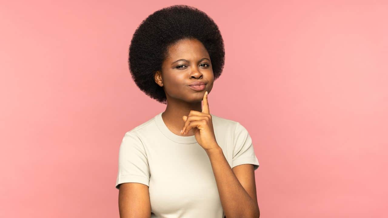 Mulher negra com a mão na bochecha pensativa, com camiseta branca e parede de fundo rosa